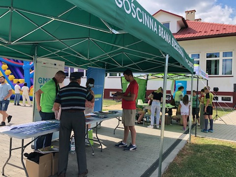 Tłum ludzi pod zielonym namiotem "Gościnna Brama Bieszczad&oacute;w". Stoliki z materiałami informacyjnymi. W tle budynek i dmuchana zjeżdżalnia.